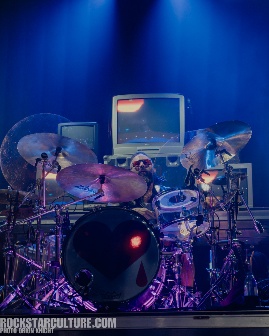 A drummer performing on stage, surrounded by drums and cymbals, with multiple vintage televisions in the background, illuminated by colorful stage lights.