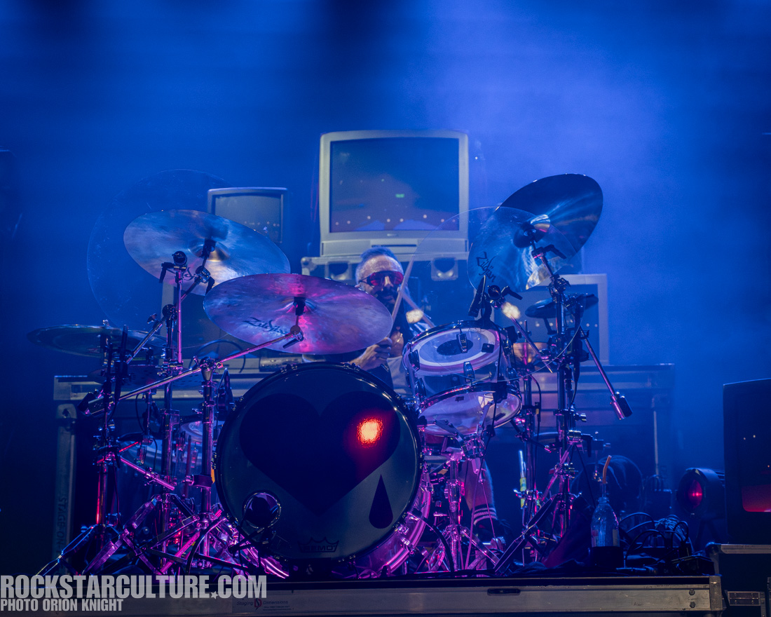 A drummer performing on stage, surrounded by colorful stage lights and retro televisions in the background.