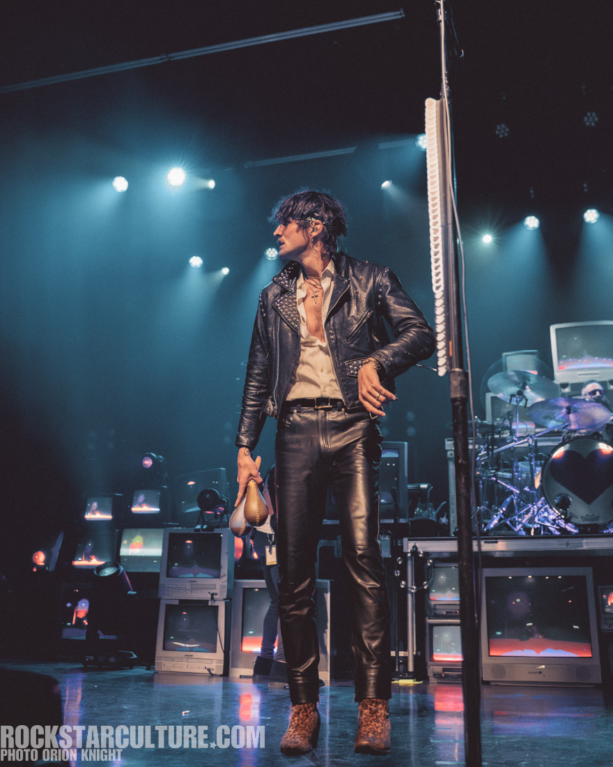A musician in a black leather jacket and tight pants holds a pair of maracas on stage, with bright lights and retro TVs in the background.
