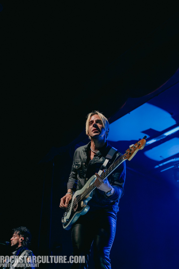 Musician playing a bass guitar on stage with focused expression, illuminated by stage lights.