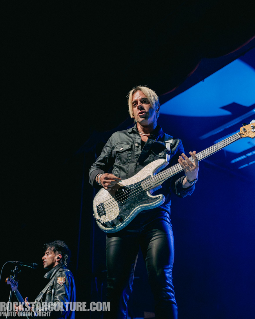 Musician performing on stage with a bass guitar, wearing a black jacket and tight pants, with a lit background.