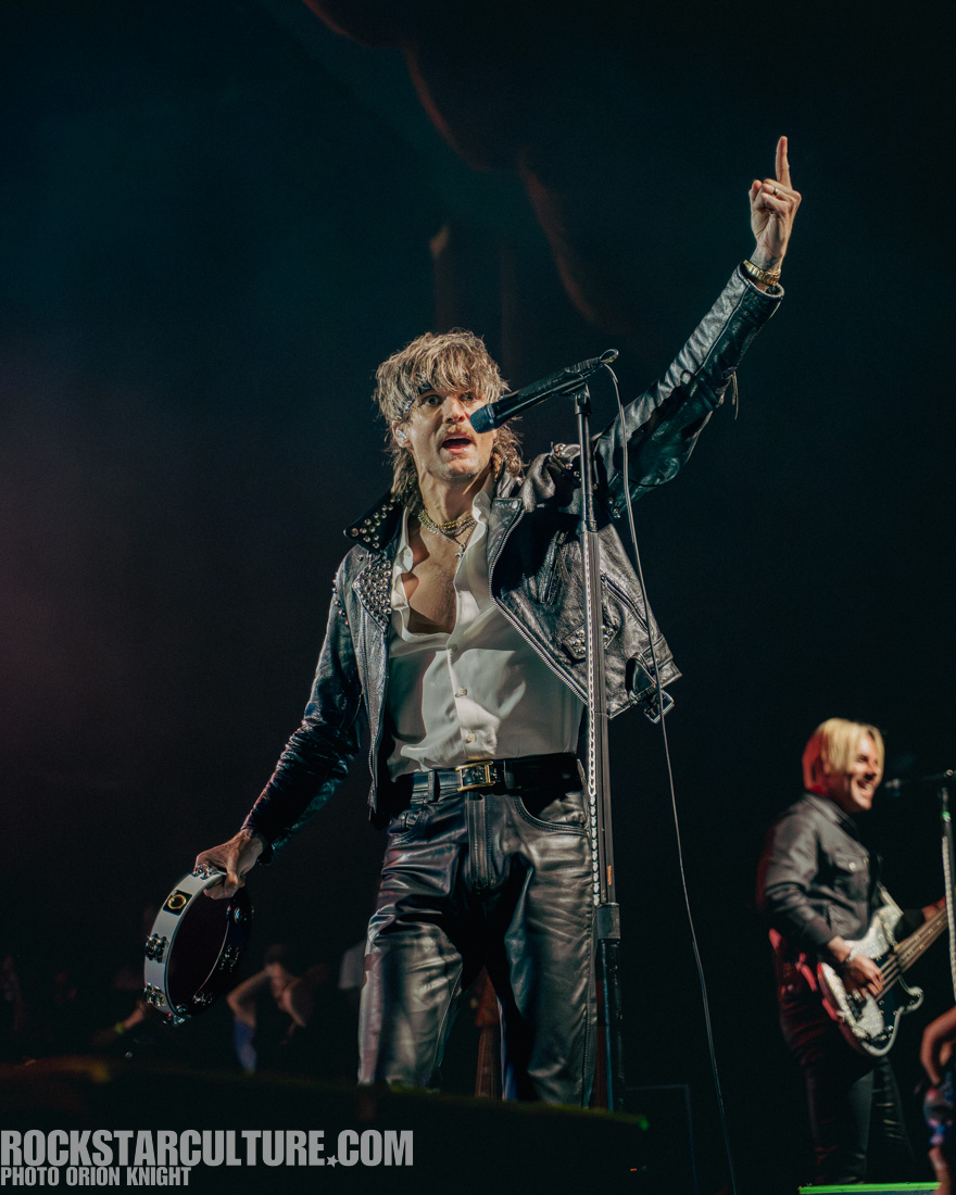 A singer in a leather jacket performs on stage, holding a tambourine and microphone, with one hand raised while a bandmate plays guitar in the background.
