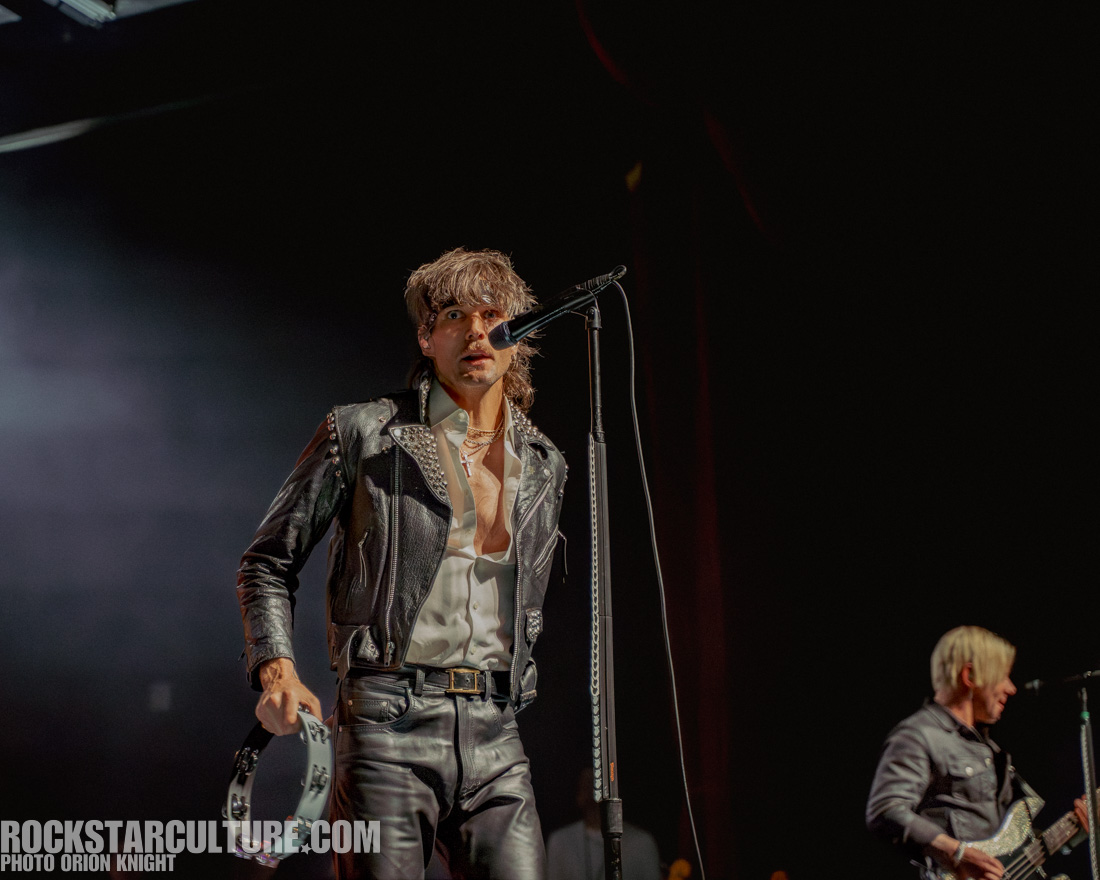 A male musician with long hair wearing a studded leather jacket and holding a tambourine, performing on stage with a microphone. Another band member is visible in the background.