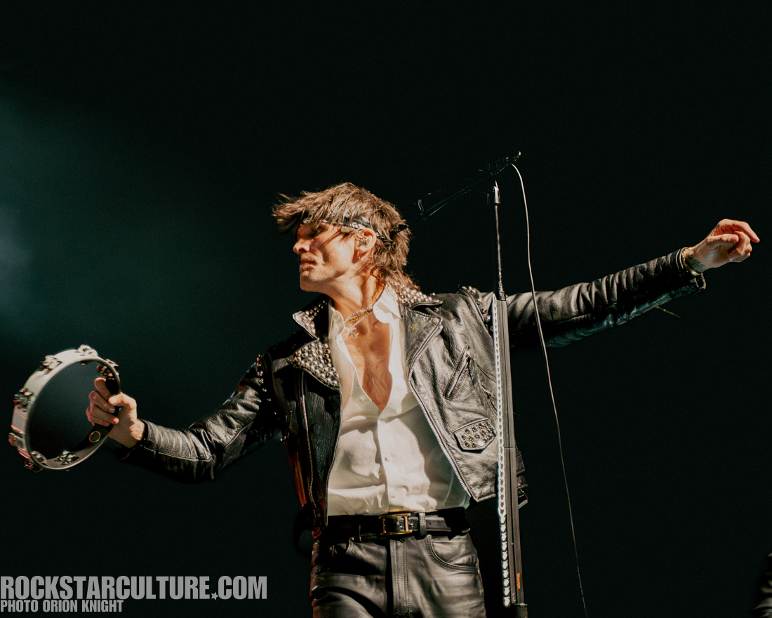 A singer performing on stage, holding a tambourine and wearing a leather jacket with studs, under dramatic lighting.