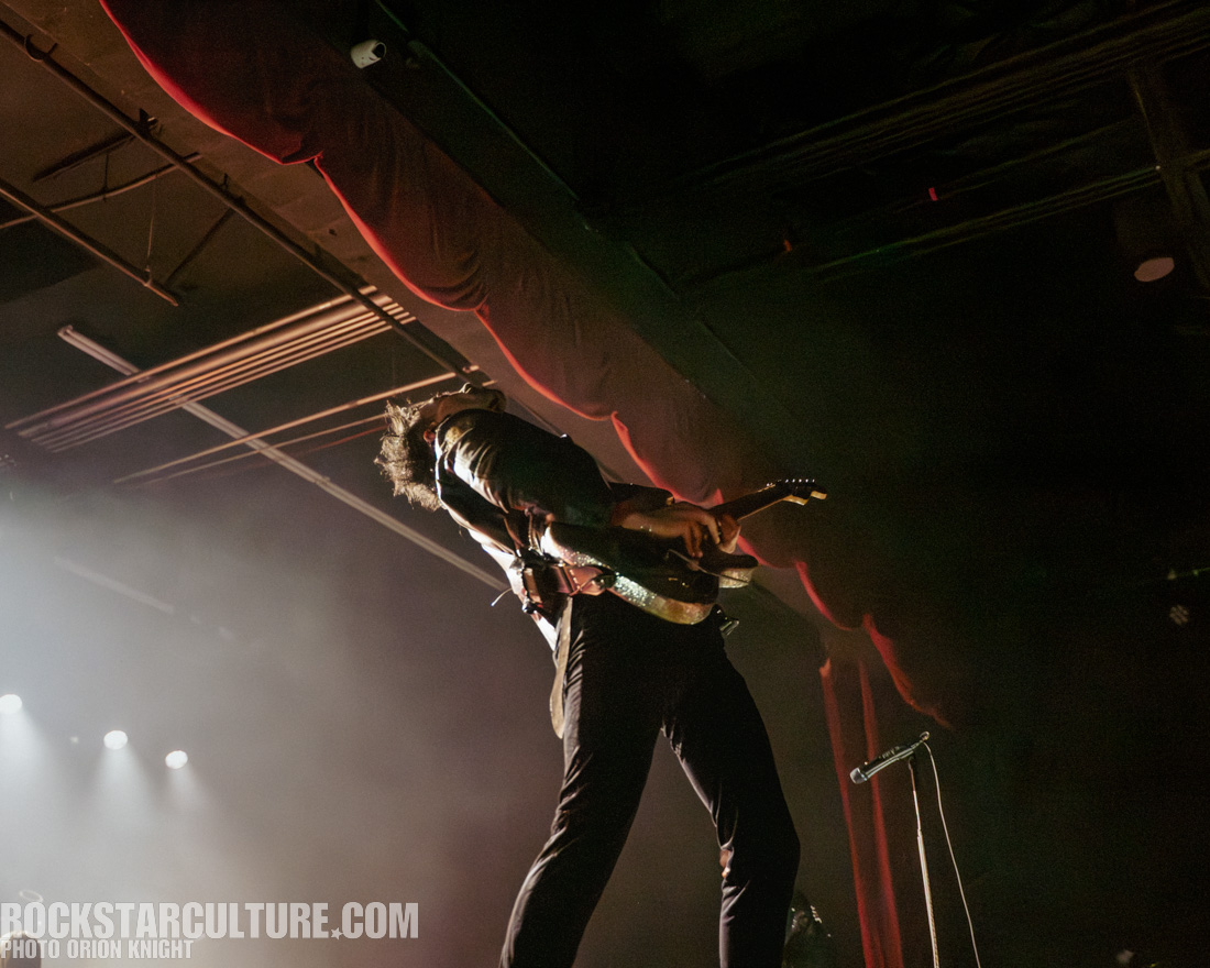 A musician energetically playing an electric guitar on stage during a live performance, with dramatic lighting and a lively atmosphere.