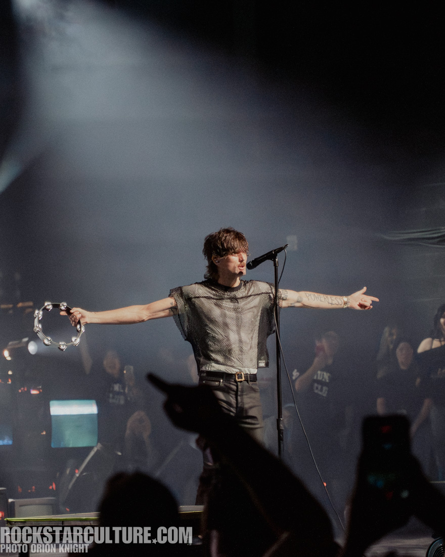 A male musician on stage performing with a tambourine in one hand and extending his arms, wearing a mesh top and black pants, with a crowd visible in the background.
