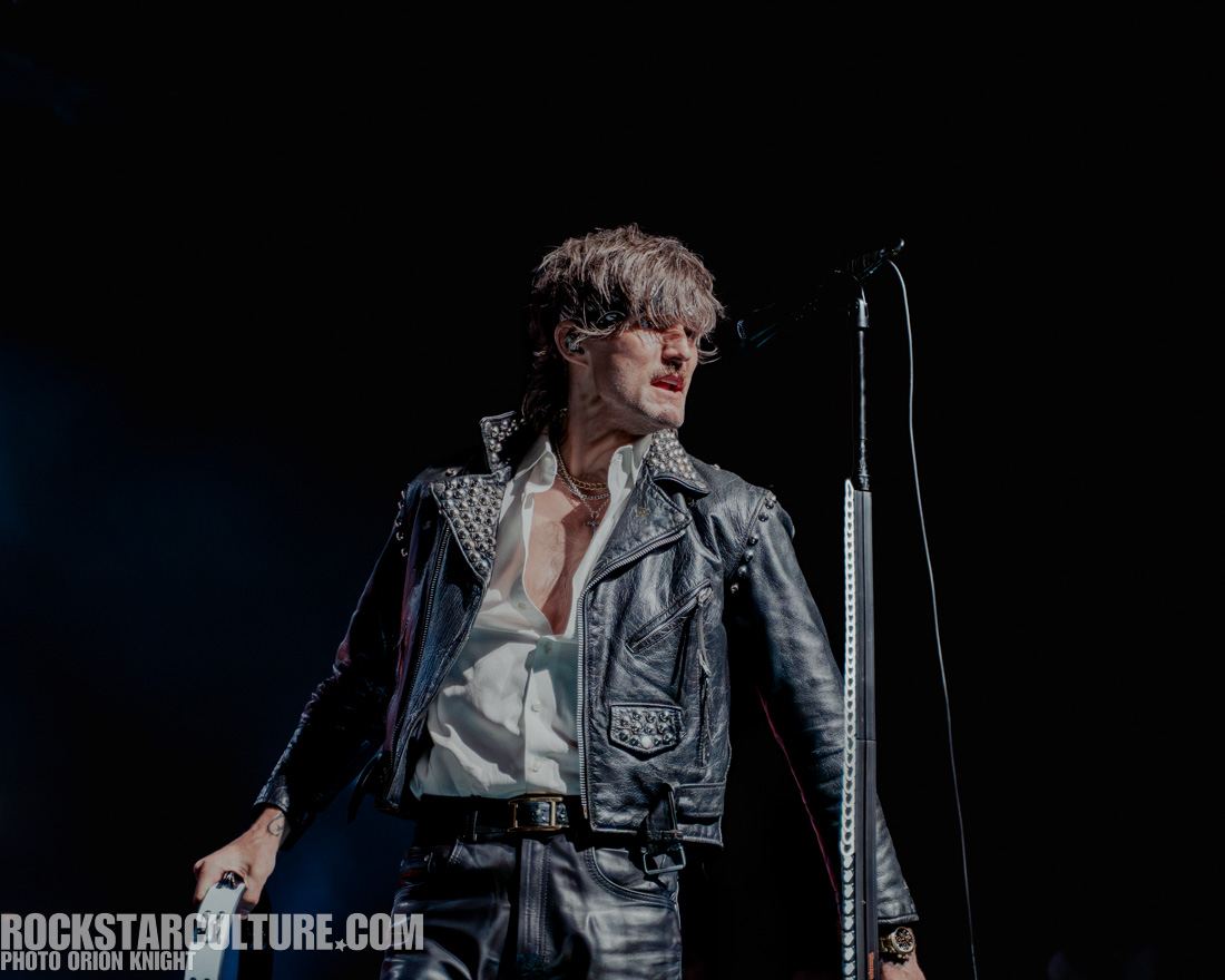 A rock musician performing on stage, wearing a studded black leather jacket and white shirt. The artist is captured mid-singing, with a microphone stand in the background and dramatic lighting.