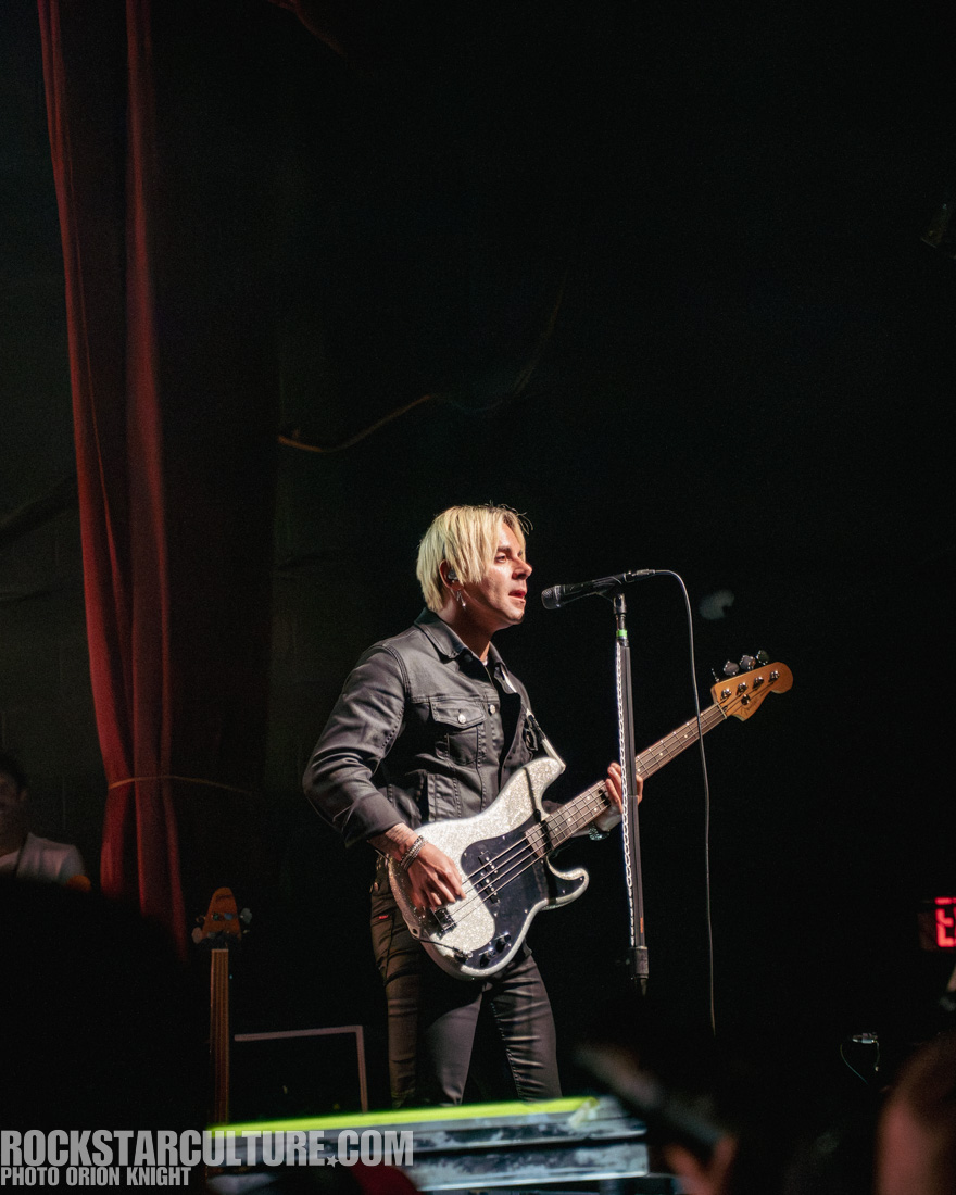 Male musician performing on stage with a bass guitar, wearing a black jacket and standing near a microphone.