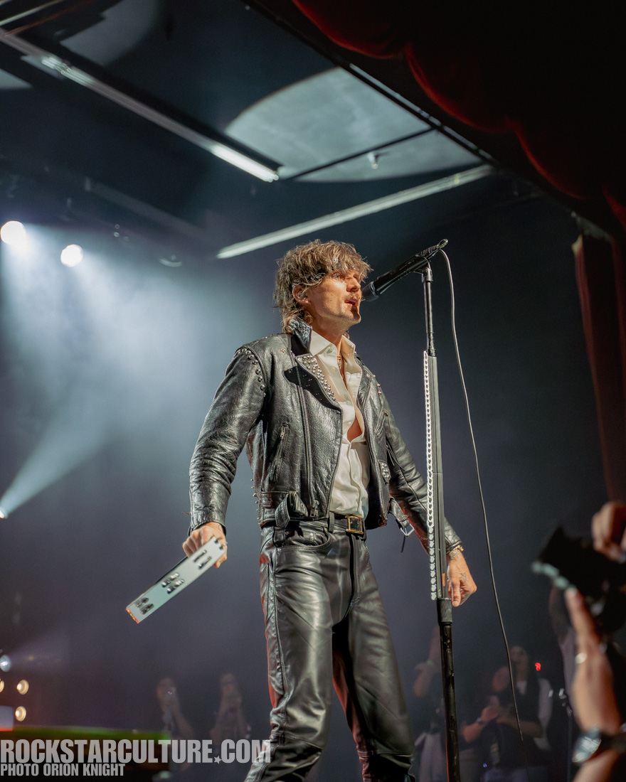 A musician performs on stage, wearing a leather jacket and black pants, with a microphone in hand and an energetic expression.