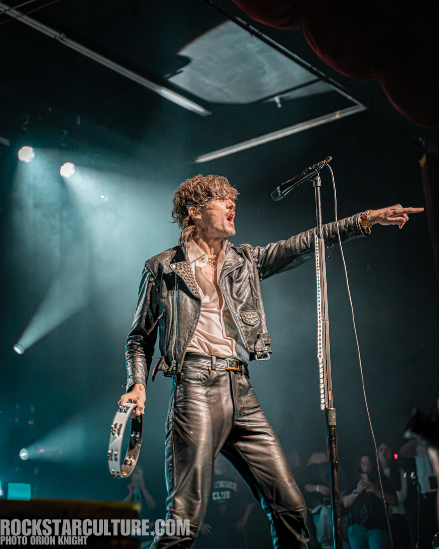 A rock musician performing on stage, wearing a leather jacket and pants, passionately gesturing with a tambourine in hand.