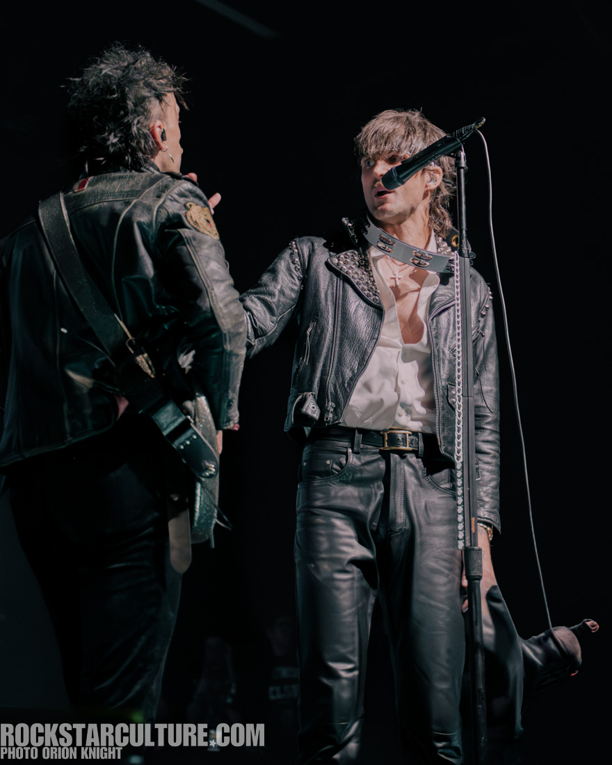 Two male rock musicians performing on stage, one with a microphone and the other playing guitar, both wearing leather jackets.