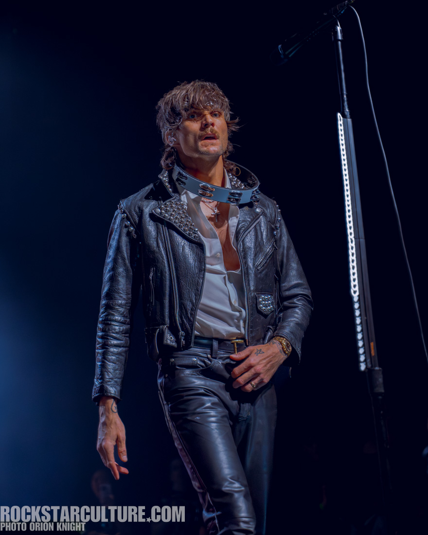 A male singer wearing a leather jacket with studs, a white shirt, and black pants performs on stage, looking intently at the audience.
