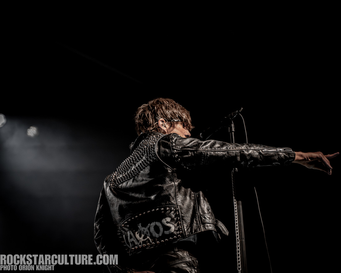 A musician in a leather jacket with studded detailing stands on stage, pointing while performing in front of a dark background.