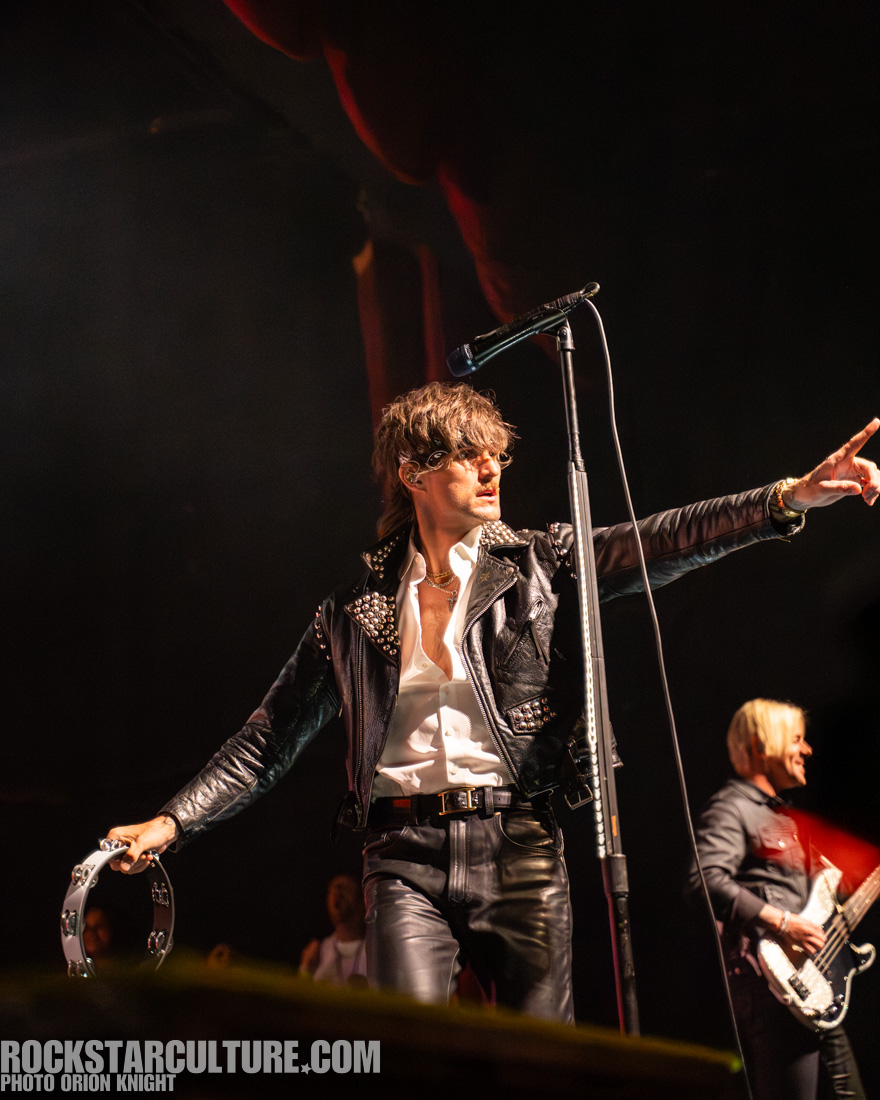 A singer wearing a black leather jacket with studs and sunglasses, holding a tambourine and pointing while performing on stage, with a band member playing bass in the background.