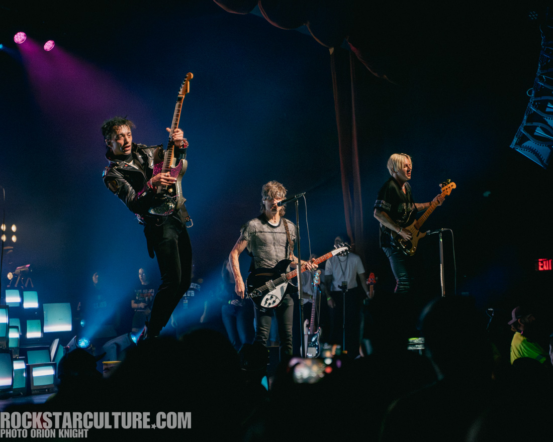 A rock band performing live on stage with vibrant stage lighting. One guitarist is jumping in mid-air while playing, and two other band members are playing guitars and engaging with the audience.