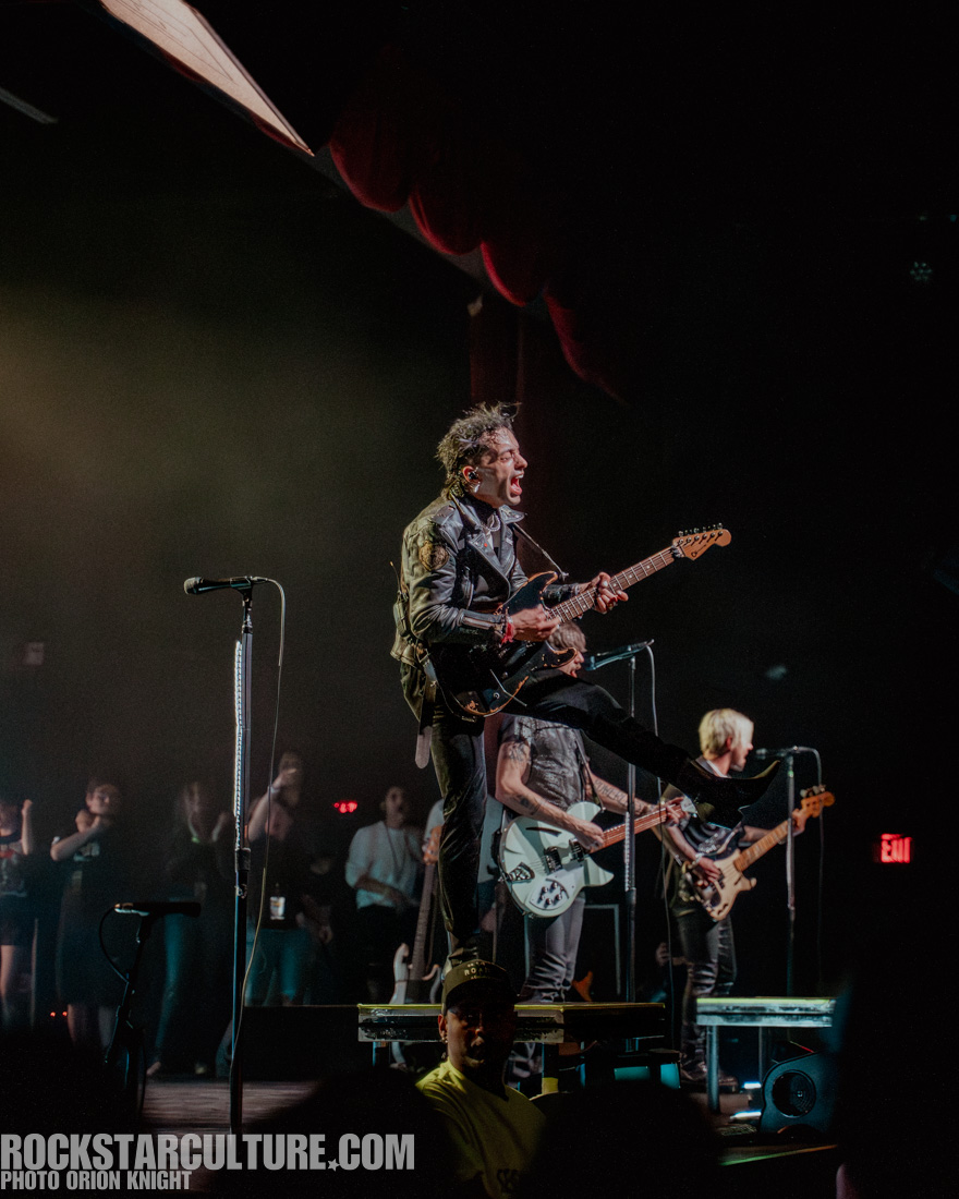 A lead guitarist in a leather jacket passionately performing on stage, with one leg raised and singing into the microphone, while a band performs behind him.