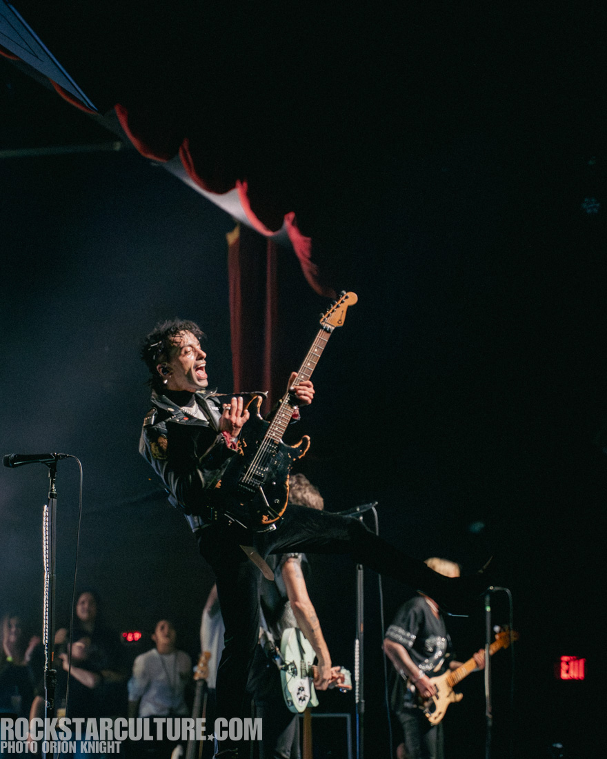 A singer performs energetically on stage with an electric guitar, dressed in a leather jacket, while a band plays in the background.