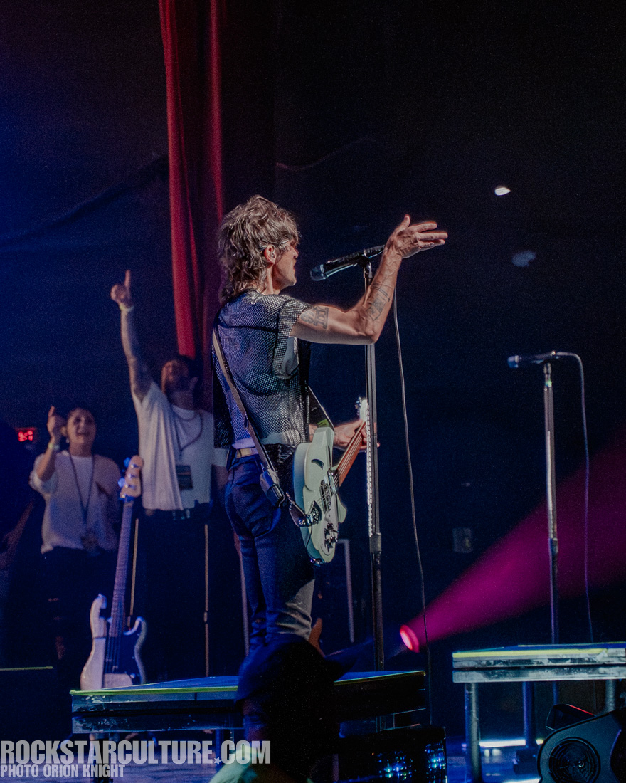 A musician performing on stage, facing away from the audience, gesturing with one hand while holding a microphone with the other. In the background, cheering fans can be seen.