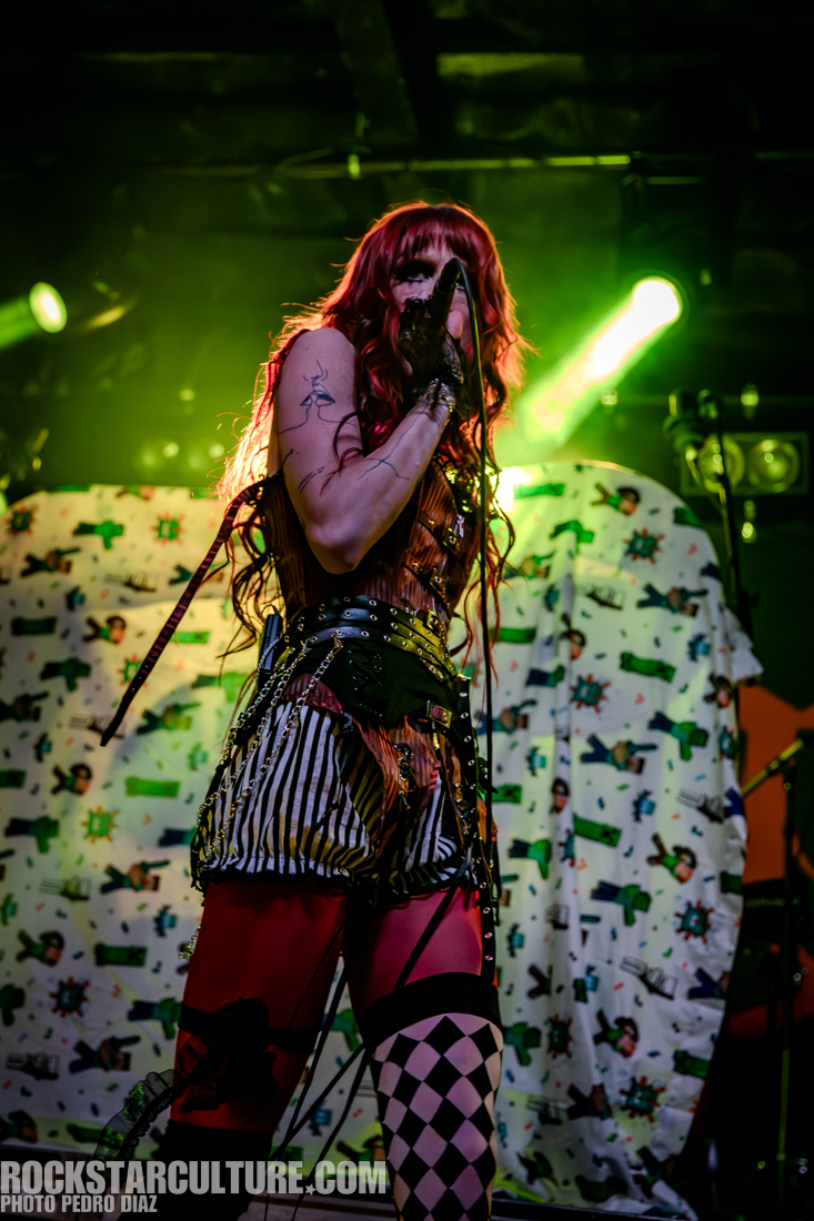 A performer with long red hair stands on stage in a colorful, eclectic outfit while singing into a microphone. The background features a patterned backdrop with various designs.