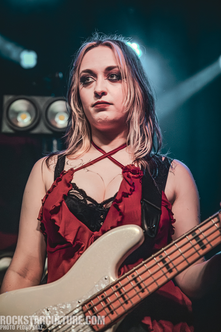 A female musician playing a bass guitar on stage, wearing a red top with black lace details and looking focused.