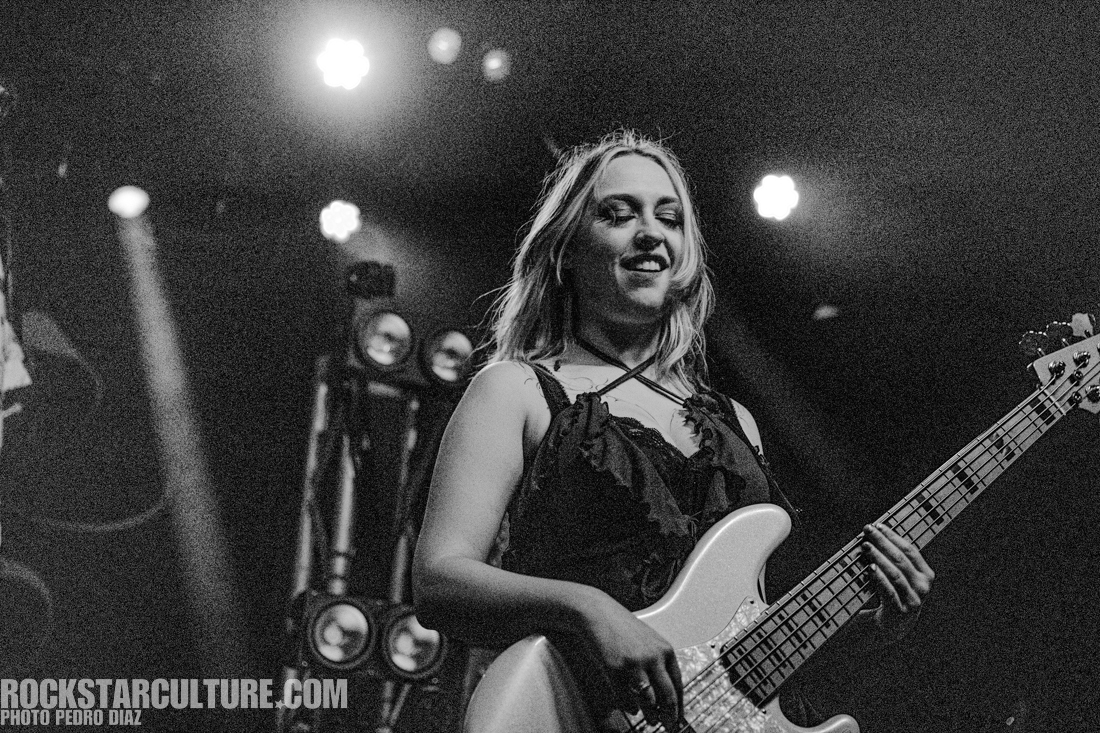 A musician playing an electric bass guitar on stage, smiling, with stage lights in the background.