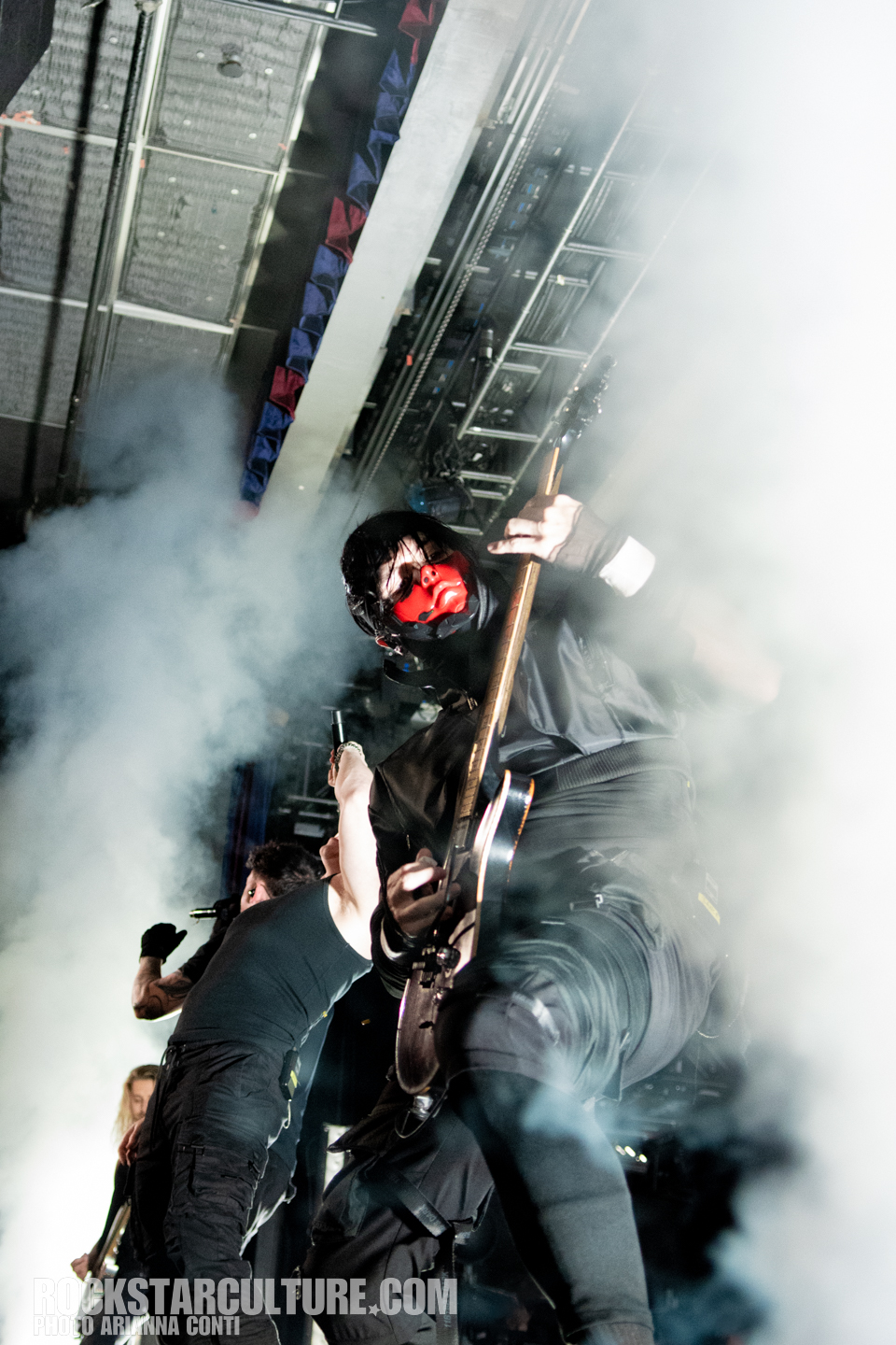 A musician wearing a red mask and black clothing plays an electric guitar on stage, surrounded by smoke, while another performer sings in the background.