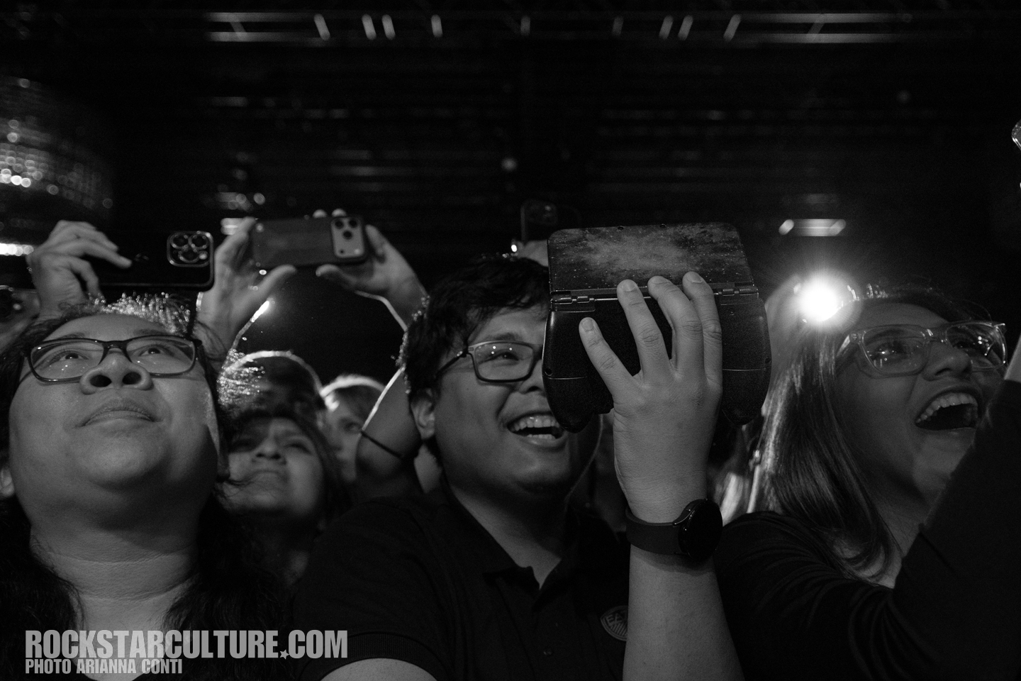 Crowd of enthusiastic concertgoers, some holding phones and a device, looking up and enjoying the performance in a lively atmosphere.
