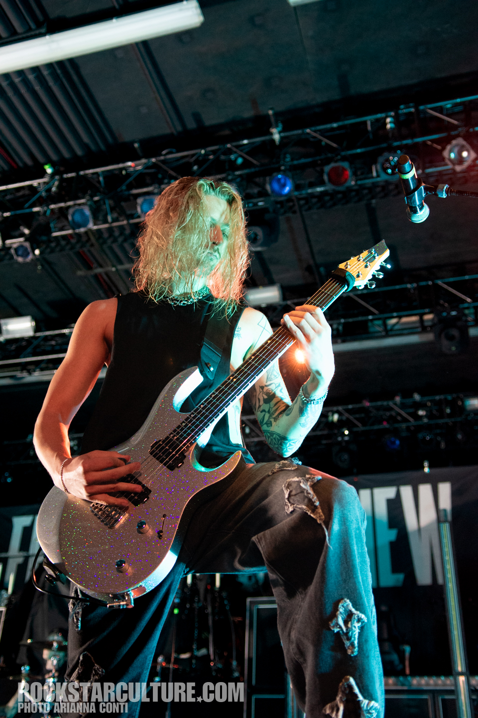 A guitarist performing live on stage with a sparkling silver guitar, wearing a sleeveless black shirt and distressed jeans, illuminated by stage lights.