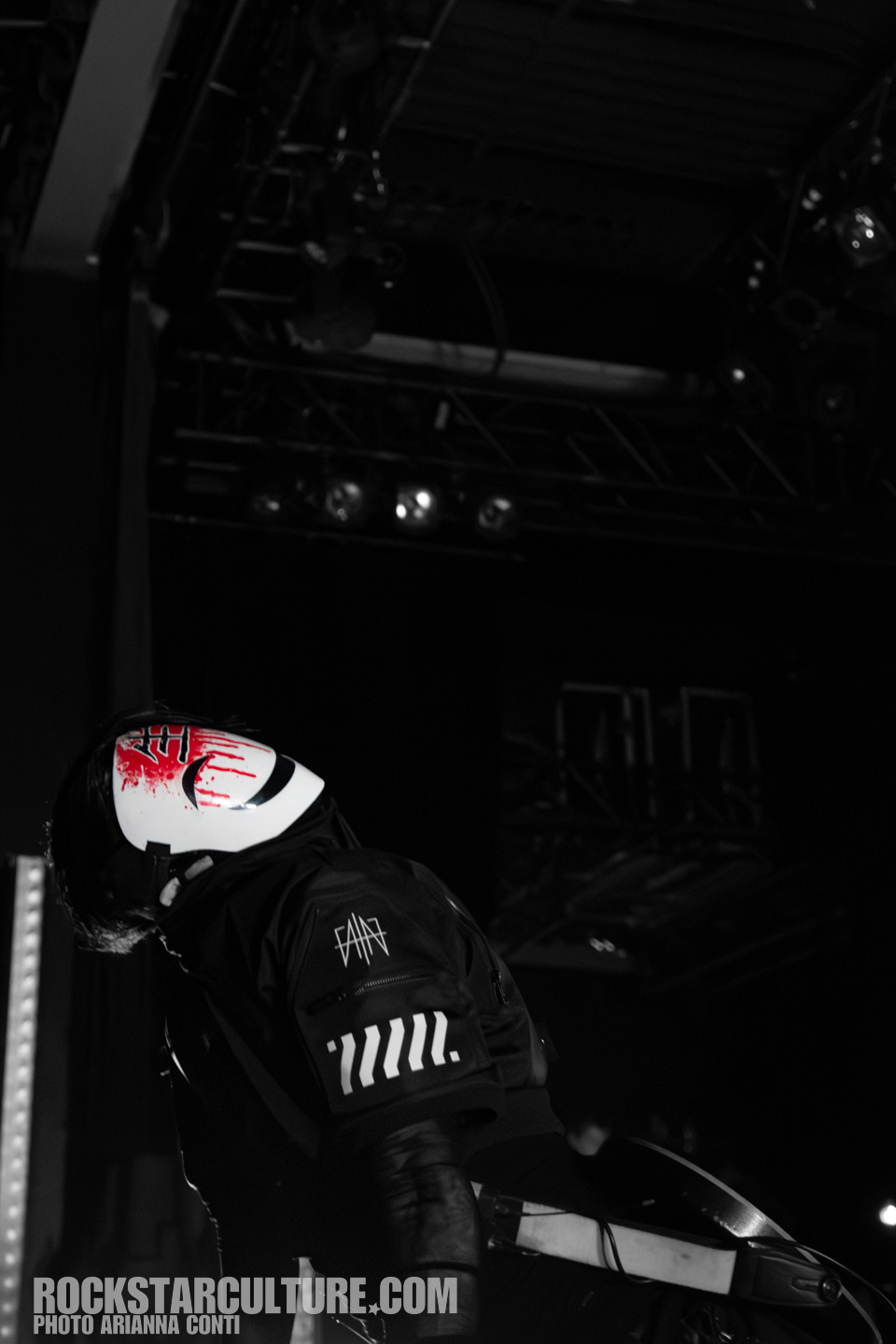 A performer wearing a distinctive white mask with red accents, dressed in a black jacket, leans forward on stage in a dramatic pose, with lighting equipment and dark surroundings visible in the background.