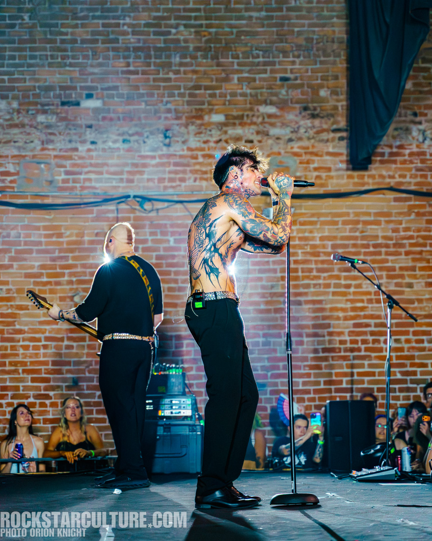 A live performance featuring a male vocalist with tattoos singing into a microphone, while a guitarist plays in the background. The scene is set in an industrial venue with a brick wall, and the audience is visible in the foreground.