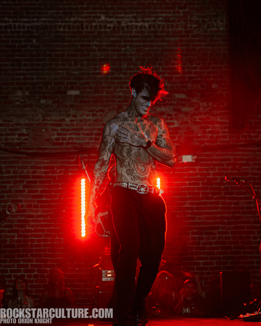 A male performer with extensive tattoos stands on stage, showcasing his physique while smiling. The background features a red lighting effect against a brick wall.