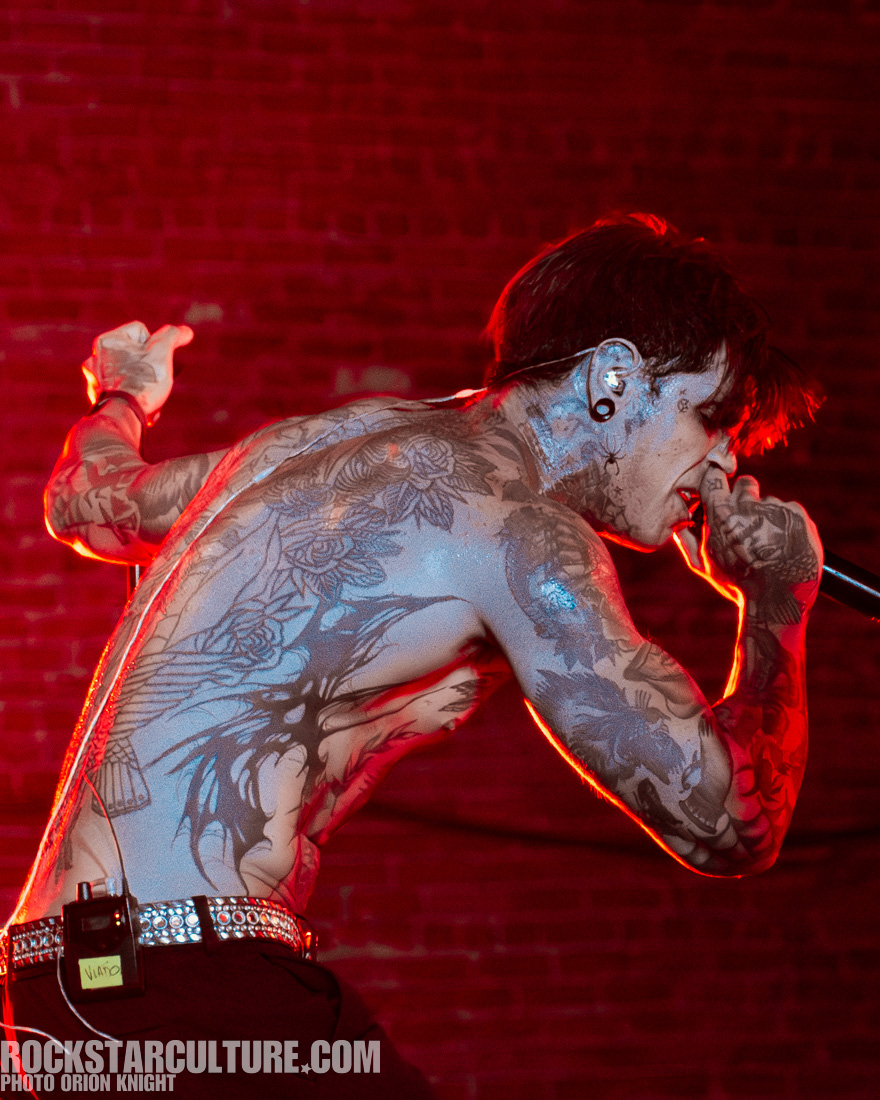 A tattooed male performer singing passionately on stage, illuminated by red lighting.