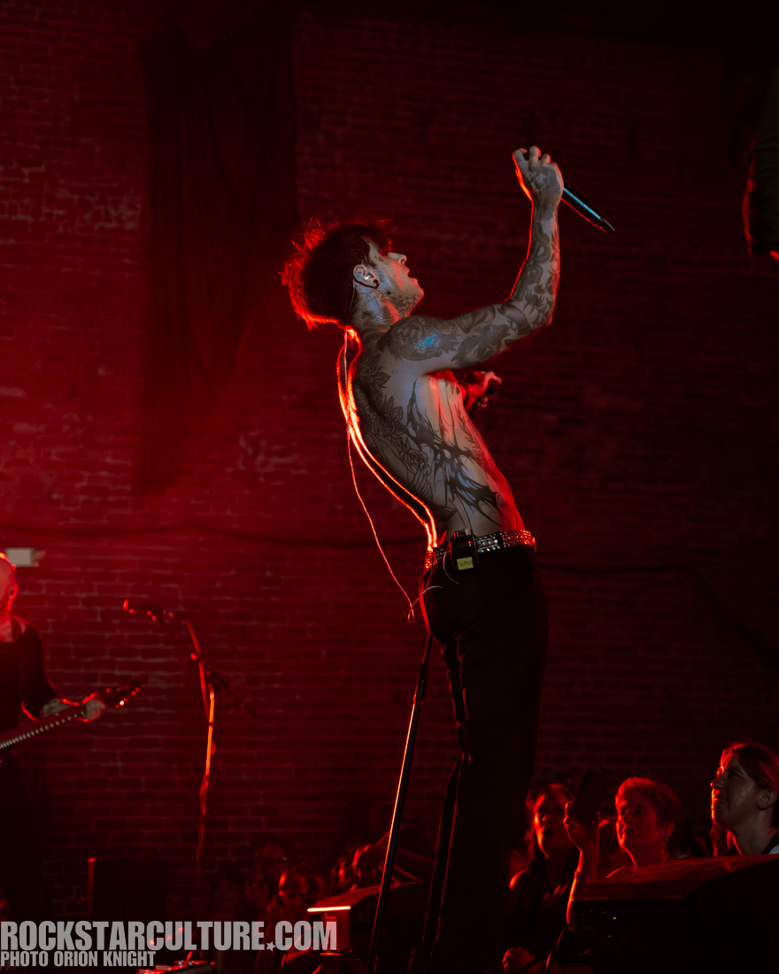 A shirtless male singer passionately performing on stage with tattoos visible and dramatic red lighting in the background.