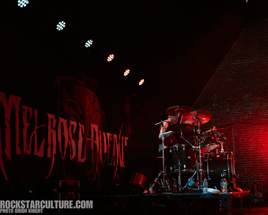 A drummer performing on stage in front of a backdrop featuring the text 'Melrose Avenue'. The venue has dramatic lighting with a mix of red and white lights, creating an energetic atmosphere.