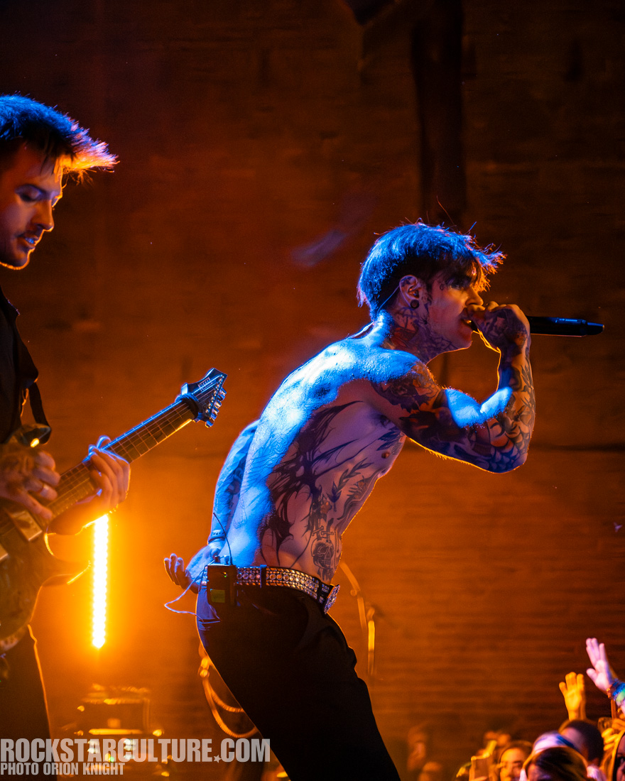 A tattooed male singer performing on stage with a guitarist in the background, under dramatic lighting in a lively concert setting.