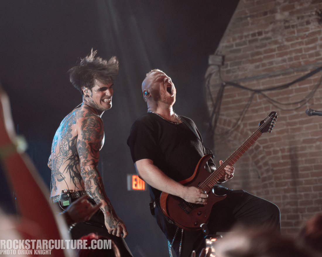 A male musician with tattoos is smiling and performing on stage alongside another male musician who is passionately playing an electric guitar.