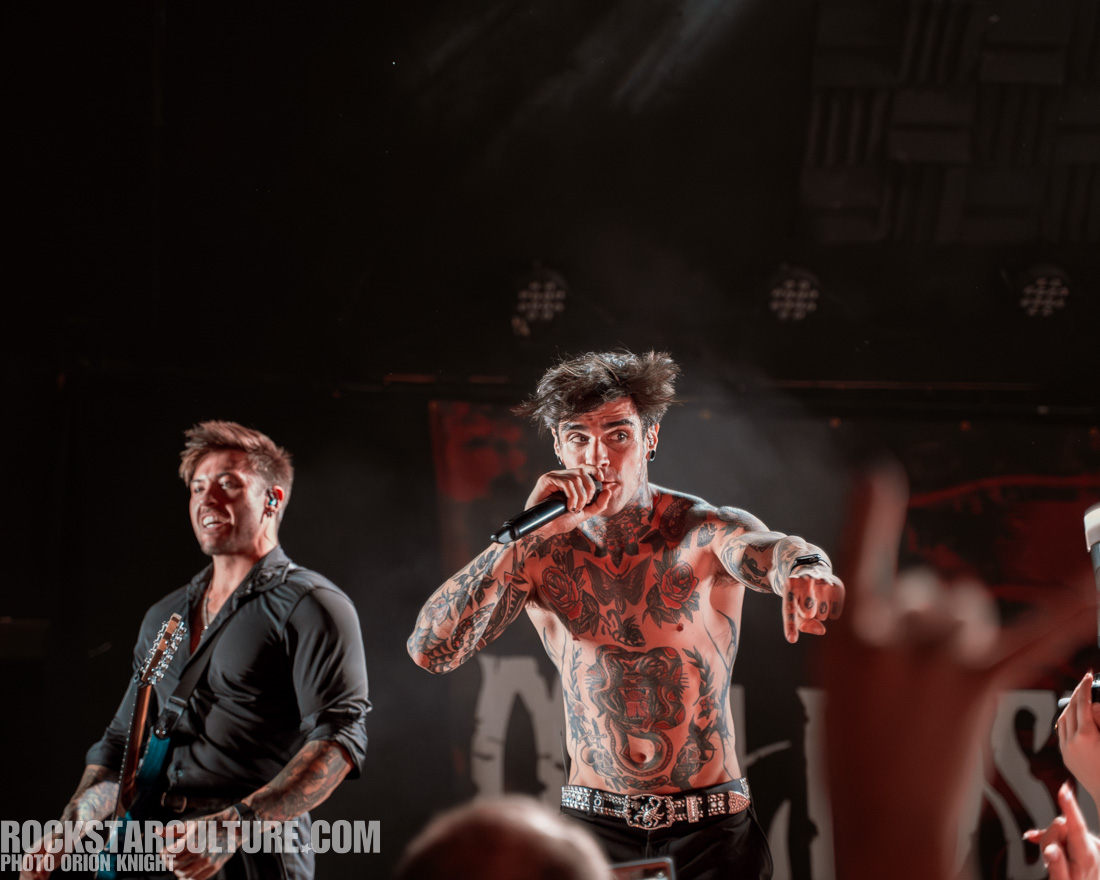 A male vocalist with tattoos performs on stage, holding a microphone and engaging with the audience. Another band member on the left plays guitar, while fans cheer in the foreground.