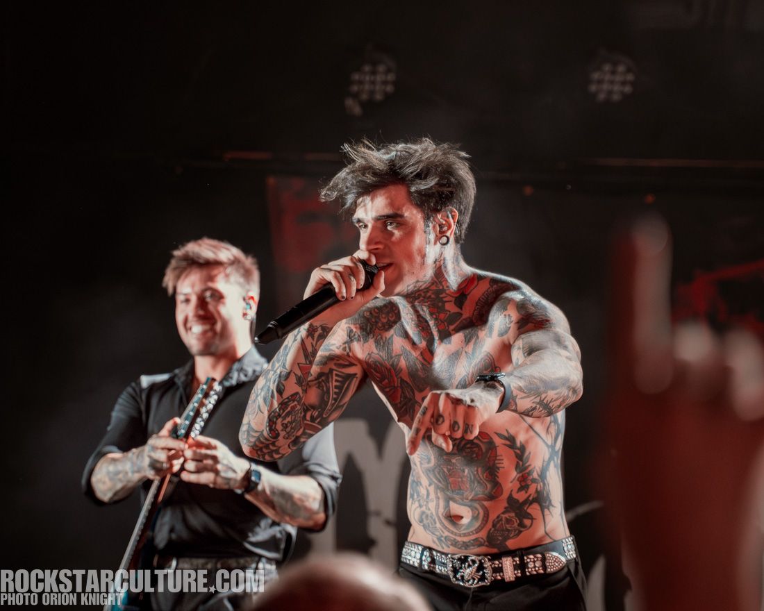 A shirtless male singer with tattoos on his torso performs on stage, holding a microphone. Another male band member with short hair smiles in the background, holding a guitar.