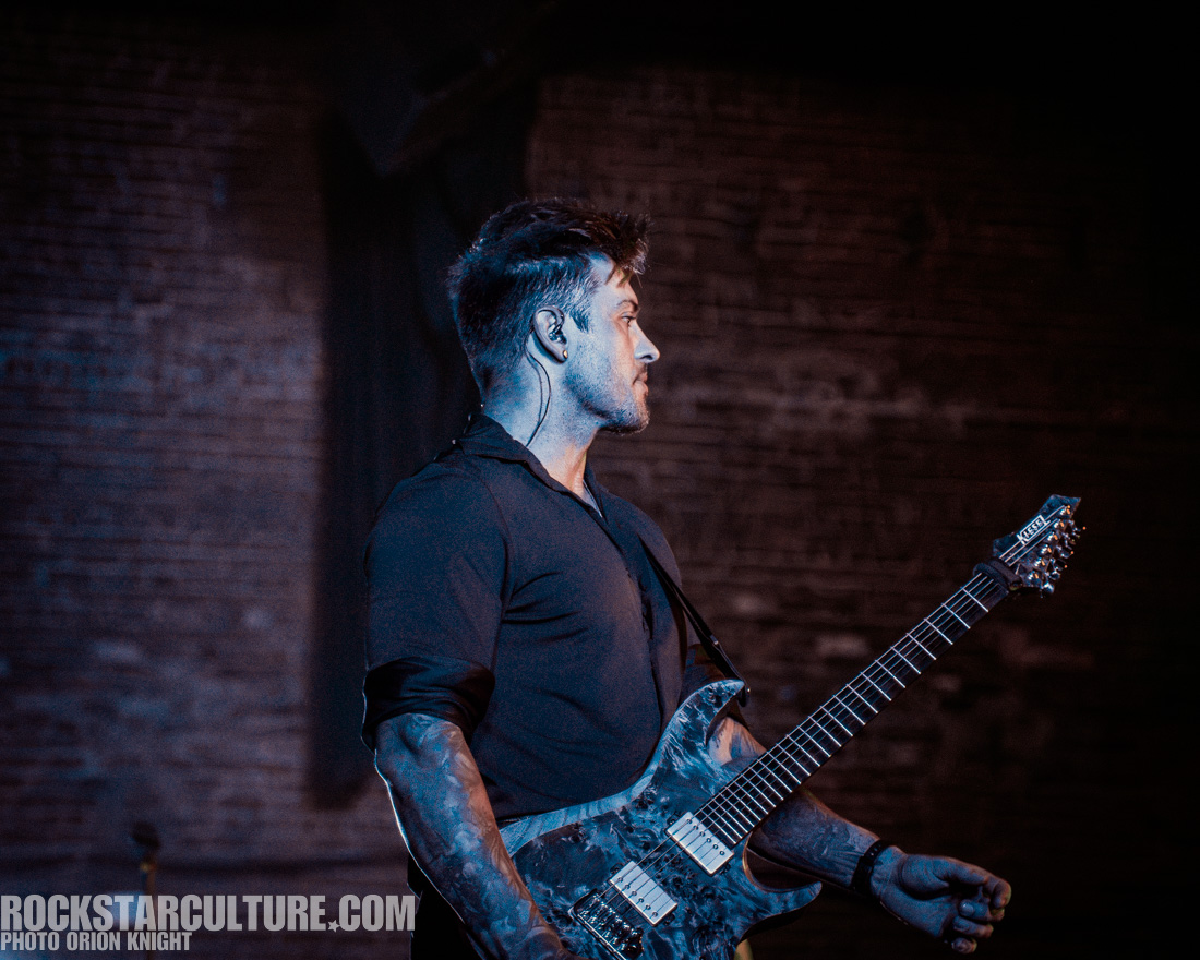 A musician on stage holding a guitar, performing in front of a brick backdrop.