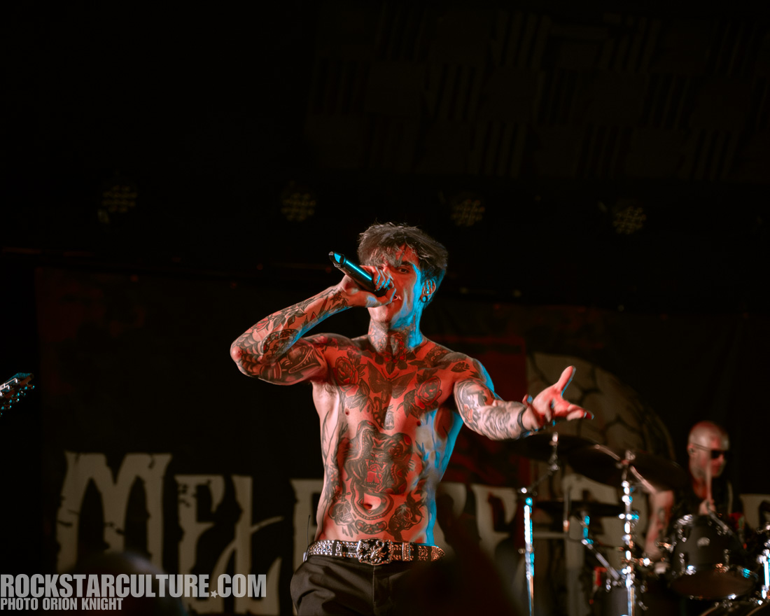 A tattooed male vocalist performing on stage with a microphone, passionately engaging with the audience, while a drummer is visible in the background.
