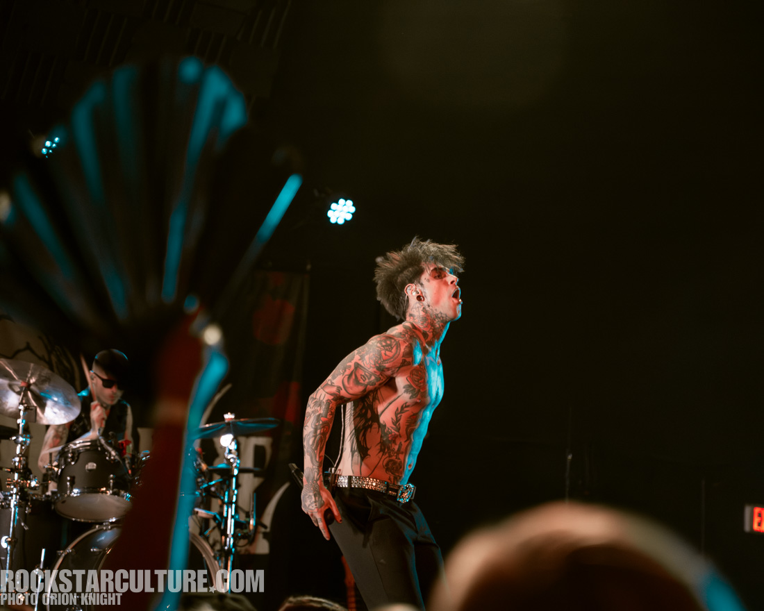 A tattooed performer on stage passionately singing, with a drummer visible in the background.