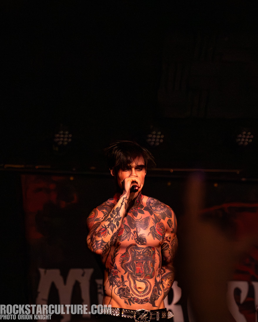 A tattooed musician performing on stage, holding a microphone, with an engaging expression, surrounded by dim lighting and a blurred audience.