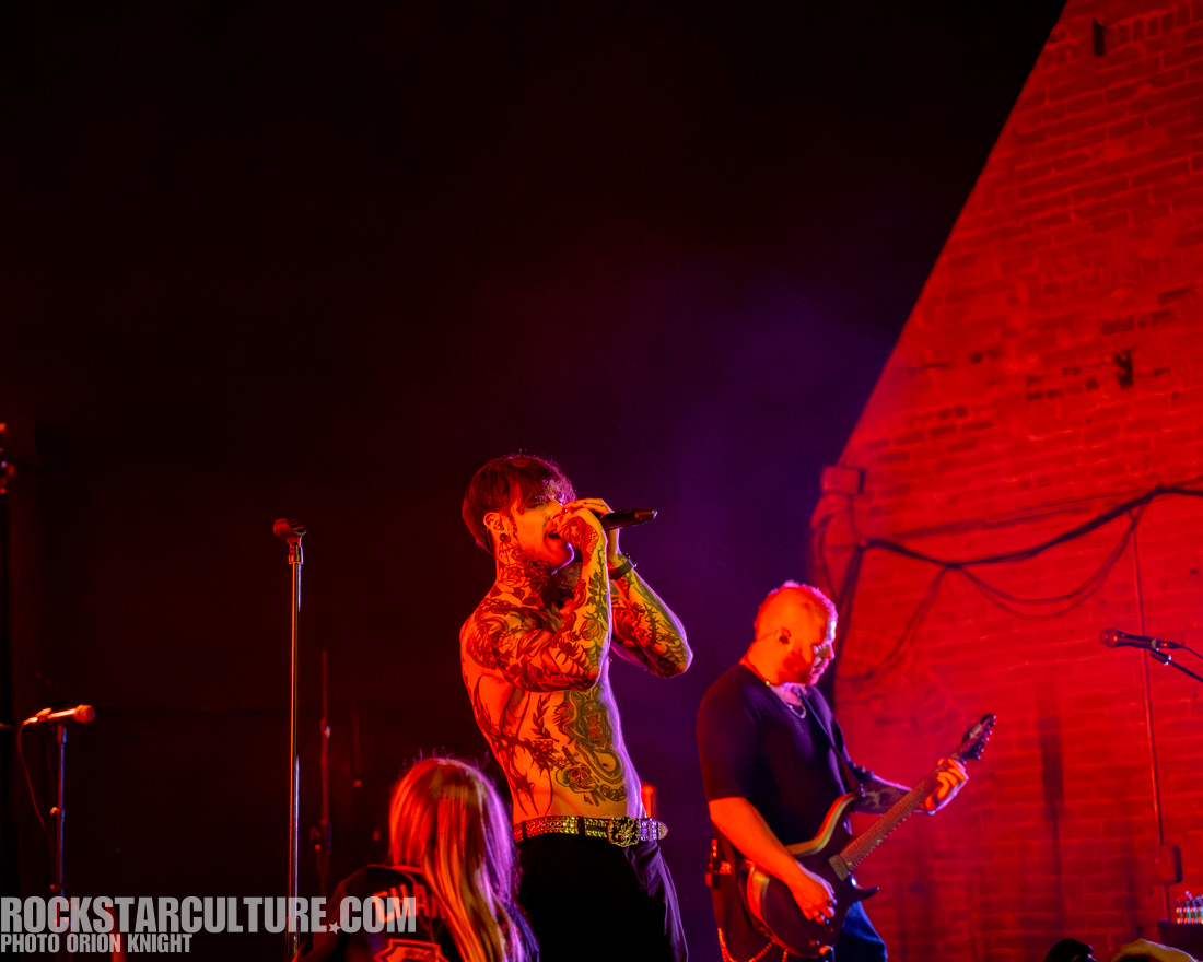 A male singer with tattoos performing on stage, holding a microphone, while another musician plays guitar in the background. The stage is illuminated with vibrant lighting, creating a dynamic atmosphere.