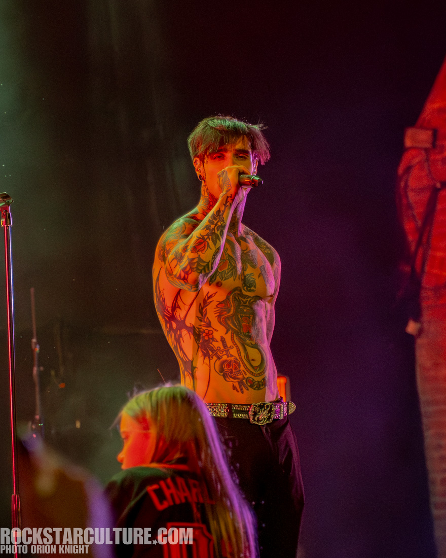 A tattooed male singer performing on stage with a microphone, surrounded by colorful lighting.