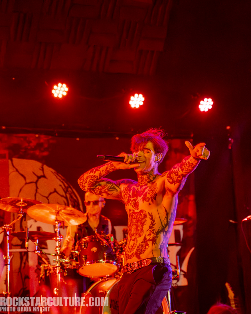 A tattooed singer performing energetically on stage with a microphone, surrounded by a drummer in the background, illuminated by red stage lights.