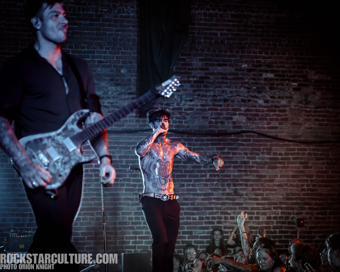A live music performance with a tattooed male singer on stage, passionately engaging with the audience. Another musician playing an electric guitar is in the background. The setting features brick walls and concert lighting.