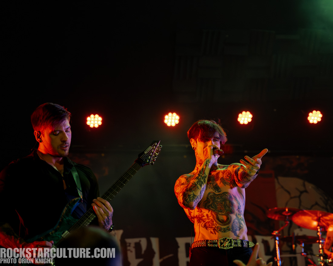 A male musician with tattoos performs on stage, holding a microphone and gesturing, while another musician plays guitar in the background, illuminated by stage lights.