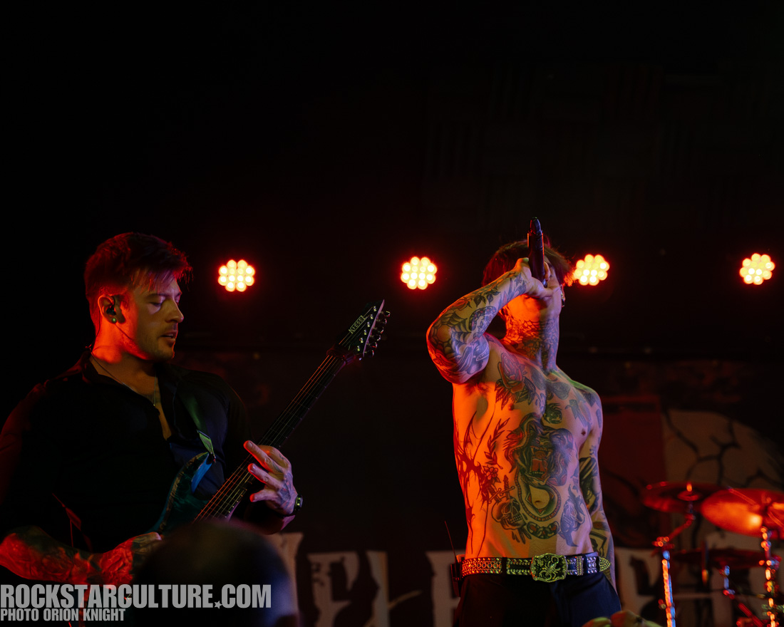 A male musician playing an electric guitar on stage, with a focus on his tattoos and intense performance. Another male performer is singing nearby, both illuminated by stage lights.