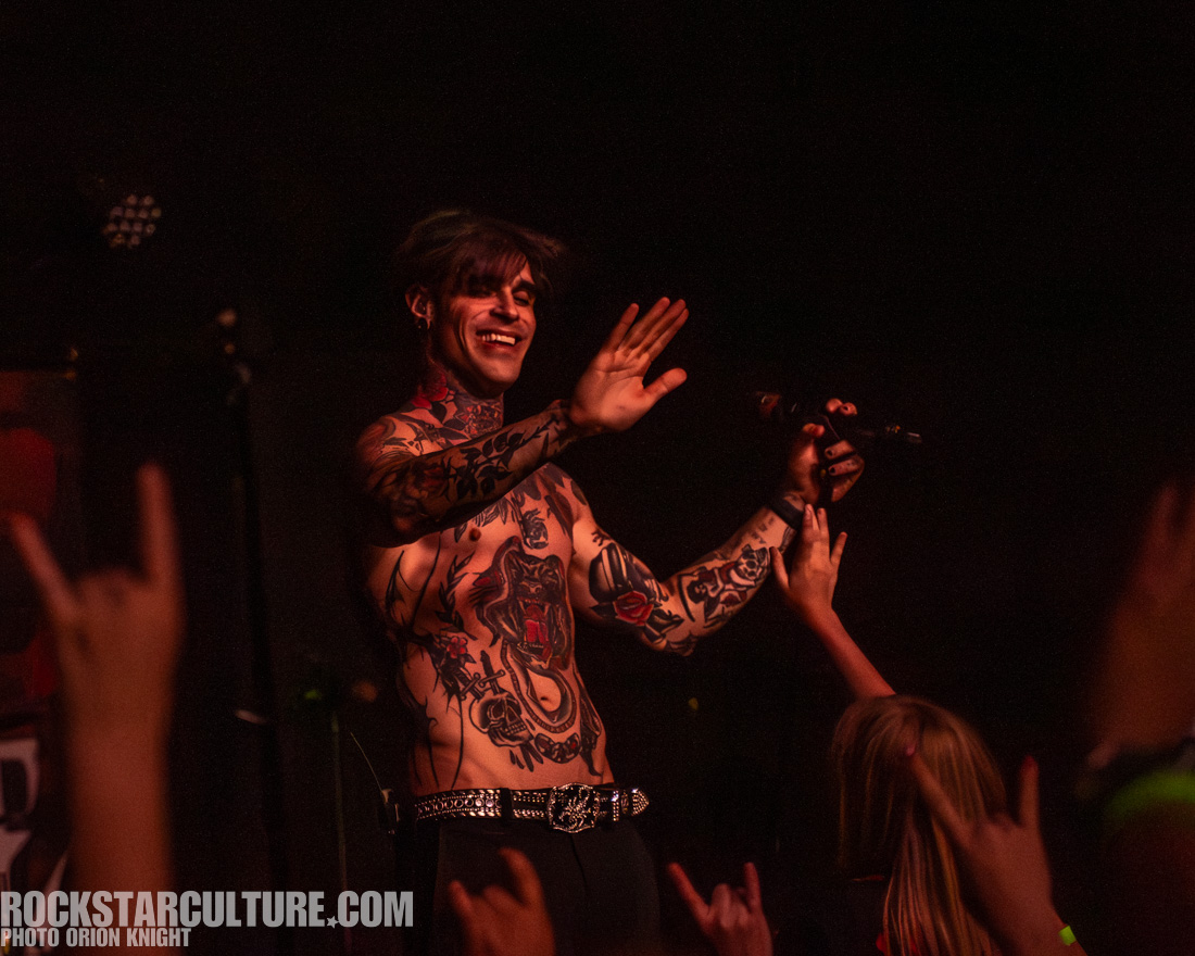 A tattooed male singer performing on stage, waving to the audience with a microphone in hand, while fans hold up hand gestures in the foreground.