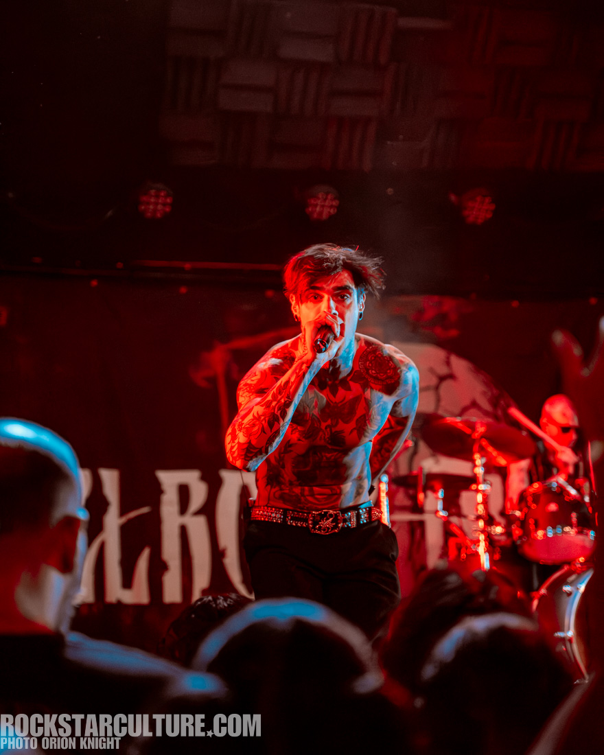A shirtless performer with tattoos passionately singing into a microphone on stage, with a drummer visible in the background and red lighting illuminating the scene.